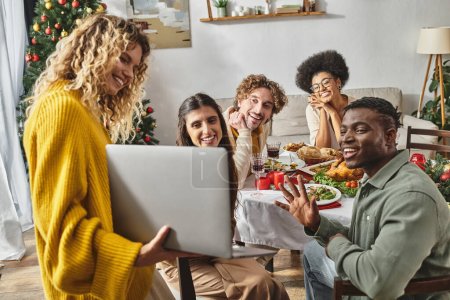 Vielvölkige Familie feiert Weihnachten beim Mittagessen jubelnd und aufrichtig lächelnd in Laptop-Kamera