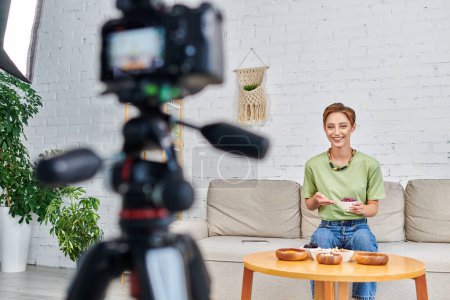 Téléchargez les photos : Blogueur vidéo végétarien souriant avec de la nourriture à base de plantes près de la caméra numérique floue dans le salon - en image libre de droit