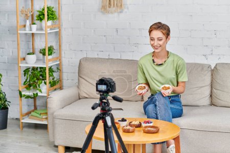 Téléchargez les photos : Femme végétarienne heureuse avec des noix présentant ensemble d'ingrédients à base de plantes pendant blog vidéo à la maison - en image libre de droit