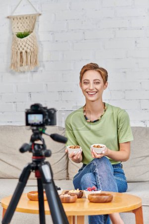 Téléchargez les photos : Joyeuse femme végétarienne tenant des bols avec des noix près de produits à base de plantes et appareil photo numérique à la maison - en image libre de droit