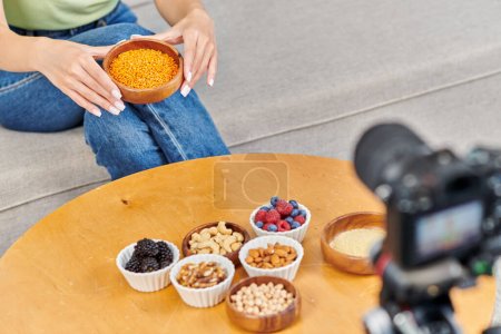 Téléchargez les photos : Femme cultivée avec bol de lentilles près de la table avec des baies fraîches et des noix devant l'appareil photo numérique - en image libre de droit