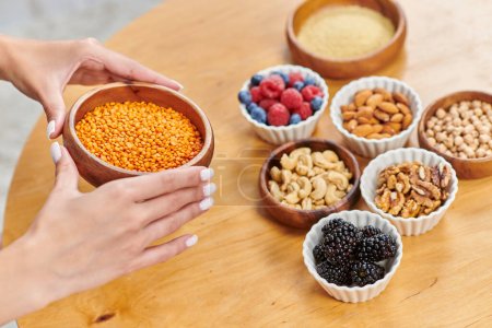 cropped woman with bowl of lentils near table with nuts and fresh berries, plant-based diets concept