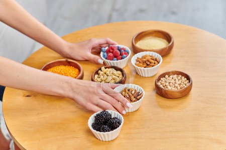 vegetarian woman placing bowls with fresh berries and various nuts with legumes on table at home