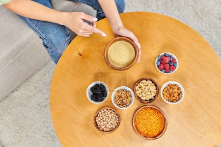 top view of vegetarian woman pointing at bowl with couscous near fresh berries and nuts with legumes