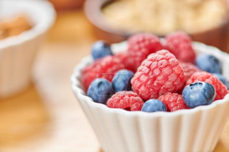 close up view of ripe raspberries and blueberries in white bowl on blurred background, vegetarianism