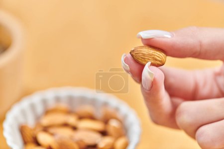close up view of female hand with almond above bowl with vegetarian food on blurred background