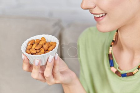 cropped view of smiley woman with white ceramic bowl of almonds, healthy vegetarian eating concept
