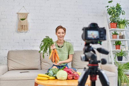 Foto de Vegetariana video blog, mujer con zanahorias frescas cerca de alimentos a base de plantas delante de la cámara digital - Imagen libre de derechos
