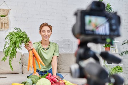 Photo for Smiling vegetarian woman with fresh carrots talking near fresh vegetables and blurred digital camera - Royalty Free Image