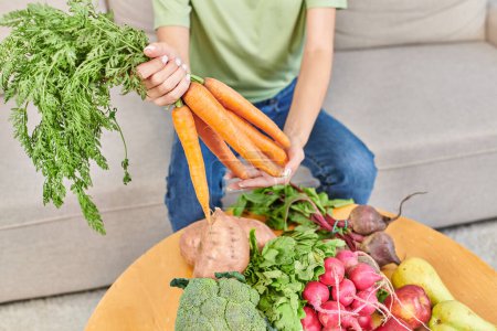 vista recortada de la mujer sosteniendo zanahorias frescas por encima de varias verduras y frutas, dietas a base de plantas