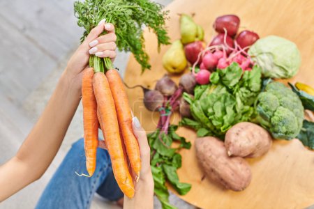 mujer vegetariana cultivada con racimo de zanahorias por encima de varias verduras y frutas, vista desde arriba