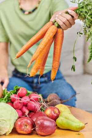 Foto de Vista parcial de la mujer con manojo de zanahorias cerca de rábano y manzanas con peras, dieta de origen vegetal - Imagen libre de derechos