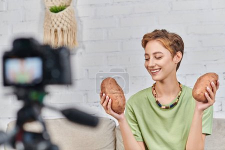 cheerful vegetarian woman holding sweet potato in front of blurred digital camera during video blog