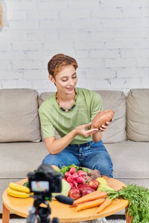 Smiley-Frau mit Süßkartoffeln in der Nähe von frischem Gemüse und Obst vor verschwommener Digitalkamera