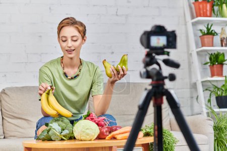 young woman with ripe bananas near various fruits and vegetables in front of blurred digital camera