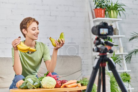 mujer vegetariana con plátanos maduros sonriendo durante el video blog cerca de verduras y frutas frescas