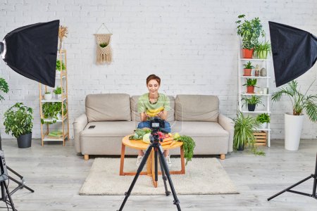 woman near plant-based food and digital camera with softbox lams in living room, vegetarian vlog