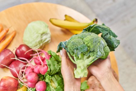 Ansicht weiblicher Hände mit frischem Brokkoli über Gemüse und Obst auf dem Tisch, pflanzliche Ernährung