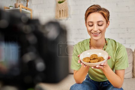 Foto de Mujer vegetariana complacida con un tazón de croquetas de verduras cerca de la cámara digital borrosa, video blog - Imagen libre de derechos