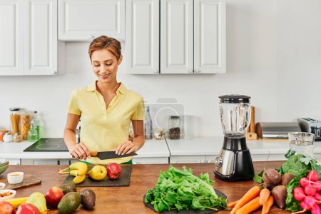 happy woman with knife near vegetables with fruits and electric blender in kitchen, plant-based diet