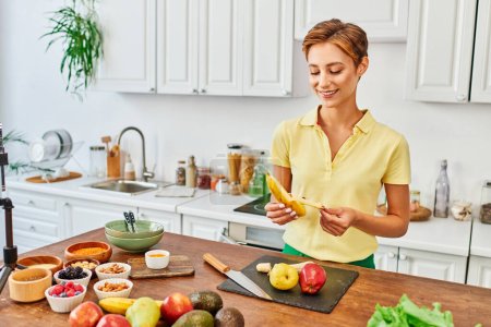 Smiley-Frau schält reife Banane in der Nähe von Obst und Gemüse auf dem Tisch in der Küche, vegetarisches Konzept