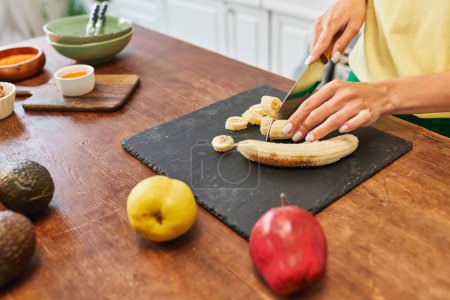 abgeschnittene Ansicht einer Frau, die reife Bananen in der Nähe von Äpfeln und Avocado schneidet, während sie eine vegetarische Mahlzeit zubereitet