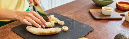 cropped view of woman cutting fresh banana near vegetarian ingredients on table at kitchen, banner