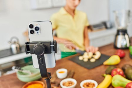 glückliche Frau schneidet Banane während Videoblog in der Nähe pflanzlicher Herkunft Zutaten und Smartphone in der Küche