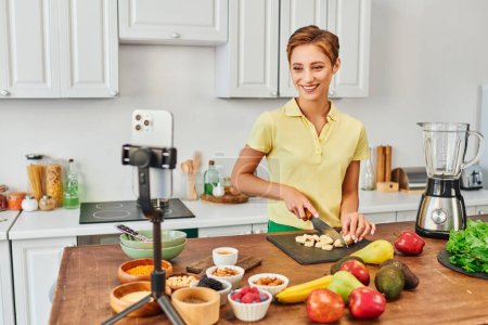 joyful woman cutting banana near plant origin food and smartphone in kitchen, vegetarian video blog