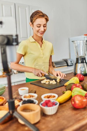 mujer encantada cortando plátano cerca de ingredientes vegetarianos y smartphone en la cocina, video blogger