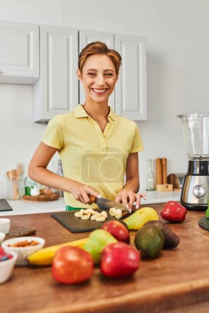 happy vegetarian woman cutting ripe banana near fresh fruits and electric blender in modern kitchen