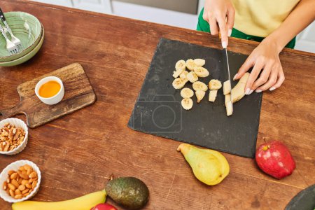 abgeschnittene Ansicht einer Frau beim Schneiden von Bananen in der Nähe von frischem Obst und Nüssen in der Küche, vegetarische Hausmannskost