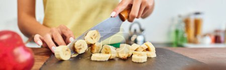 cropped view of woman cutting ripe fresh banana in kitchen, plants-based diets concept, banner