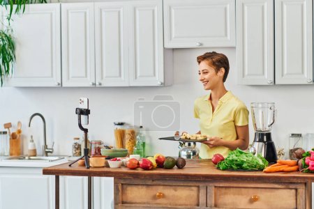 mujer feliz con plátano en rodajas en la tabla de cortar cerca de teléfonos inteligentes e ingredientes vegetarianos, vlog