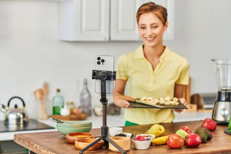 Smartphone auf Stativ neben Frau mit Schneidebrett und frischem Obst mit Gemüse in der Küche
