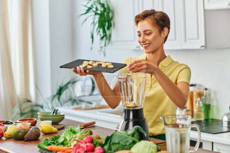 fröhliche Frau mit geschnittener Banane auf Schneidebrett neben Mixer und vegetarischen Zutaten in der Küche