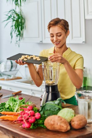 pleased vegetarian woman putting sliced banana in electric blender near fresh vegetables in kitchen