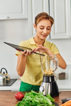 Photo for Smiling woman putting sliced banana into electric blender near fresh lettuce, plant-focused diet - Royalty Free Image