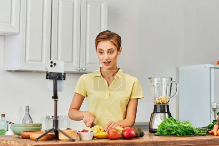mujer de corte de frutas cerca de teléfono inteligente en trípode y licuadora eléctrica en casa, vlog dieta a base de plantas