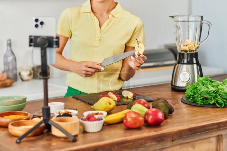 cropped view of vegetarian woman with knife and cut apple near smartphone and fruits in kitchen