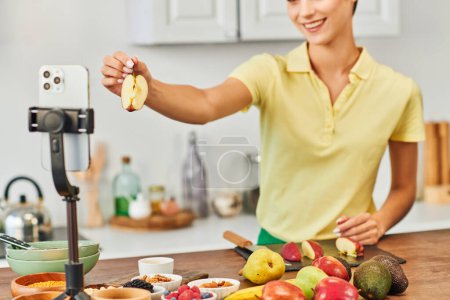 mujer sonriente recortada sosteniendo manzana cortada cerca de teléfono inteligente en el trípode y frutas frescas en la cocina