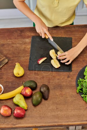 Ansicht von oben: geschnittene Frau schneidet Apfel in der Nähe von frischem Obst auf Holztisch, gesunde pflanzliche Ernährung