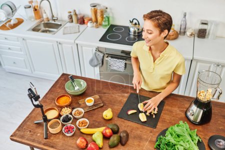 fröhliche Frau schneidet Früchte in der Nähe von Mixer und Smartphone auf Stativ, vegetarisches Videoblog