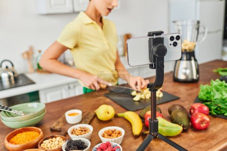 selective focus of smartphone on tripod near vegetarian video blogger cutting fruits in kitchen