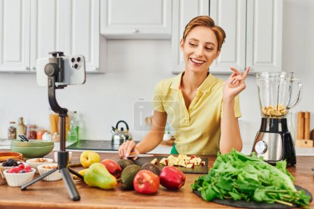fröhliche Vegetarierin schaut auf Smartphone auf Stativ in der Nähe von Obst und Mixer, vlog