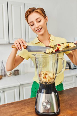 pleased woman with chopped apple on cutting board near electric blender, plant-based diets concept