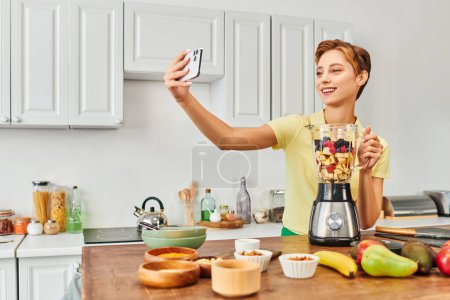 Photo for Pleased vegetarian woman taking selfie on smartphone near electric blender with fresh chopped fruits - Royalty Free Image