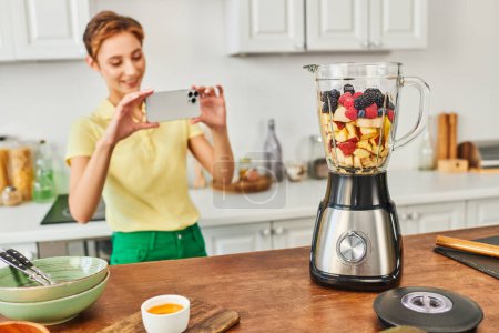 Photo for Happy woman with smartphone taking photo of electric blender with chopped fruits, vegetarian recipe - Royalty Free Image