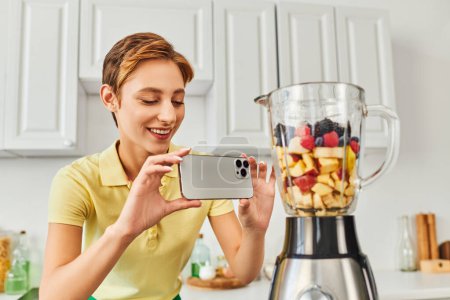 mujer positiva tomando fotos de licuadora eléctrica con frutas picadas, deliciosa dieta a base de plantas