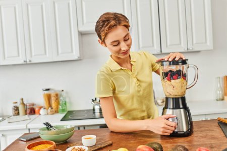 Lächelnde Frau zerkleinert frisches Obst im Mixer, während sie leckere vegetarische Smoothies in der Küche zubereitet